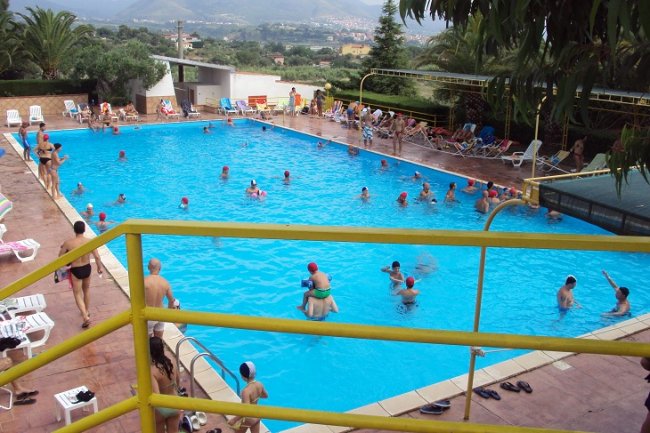 Piscina all'aperto con persone che nuotano e prendono il sole.
