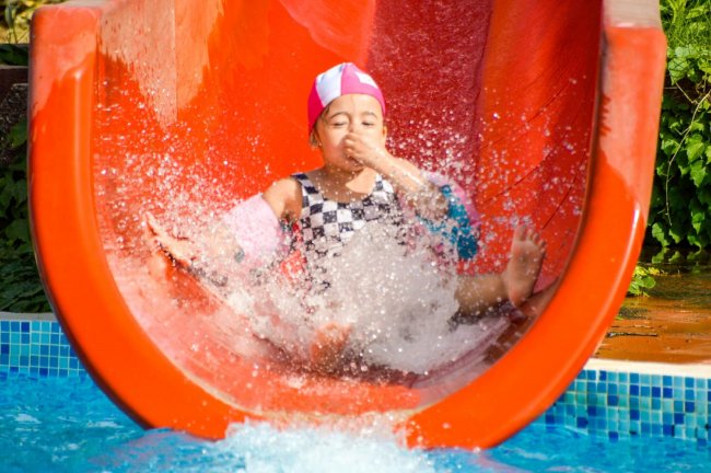 Bambino scivola divertito su uno scivolo d'acqua rosso, creando spruzzi in piscina.
