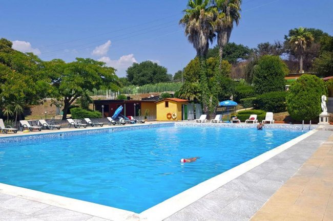 Piscina circondata da lettini e alberi sotto un cielo soleggiato.