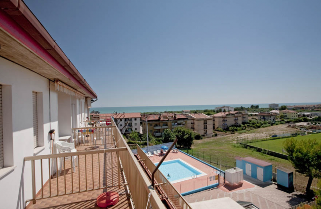 Vista di un balcone con piscina, edifici e mare sullo sfondo.