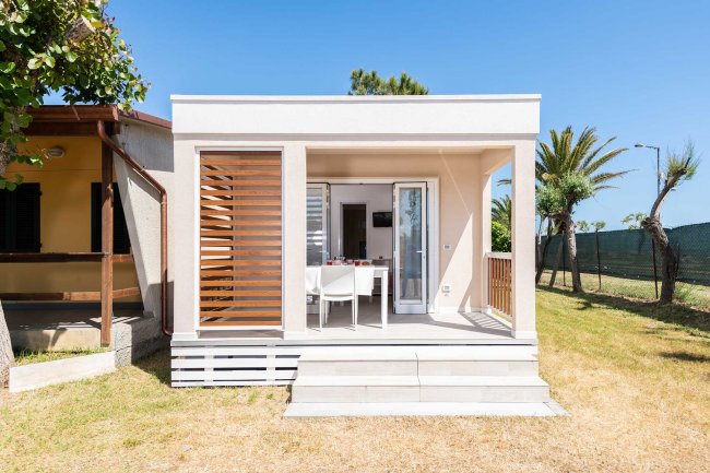 Una piccola e moderna casa prefabbricata con una veranda e sedie, circondata da un prato e alberi.