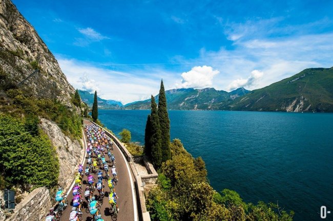 Gara ciclistica lungo un lago, montagne sullo sfondo.