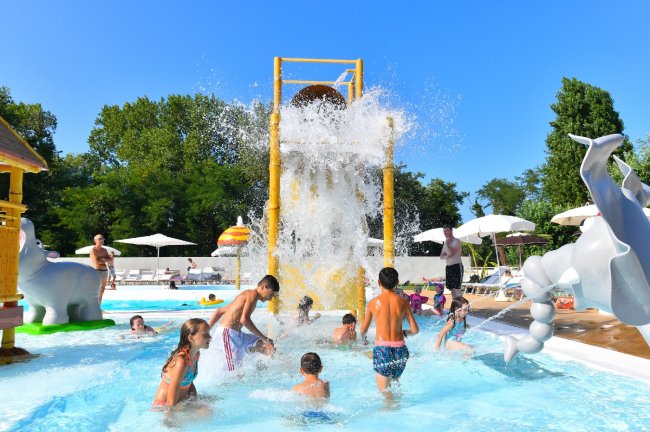 Bambini giocano sotto un grosso secchio d'acqua in una piscina all'aperto.