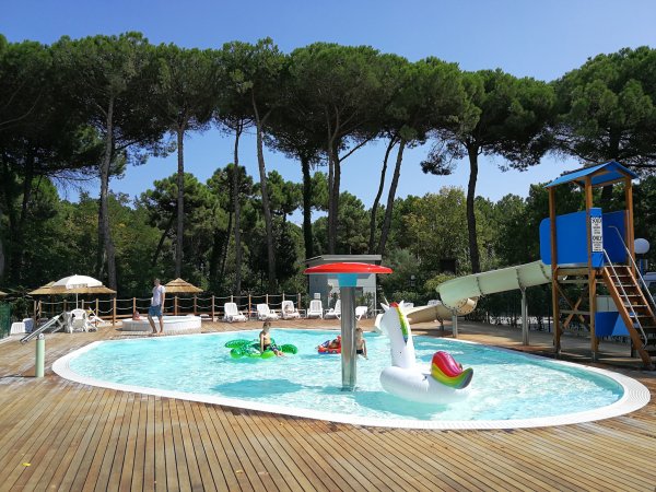 Piscina con giochi gonfiabili e scivolo circondata da alberi e lettini.