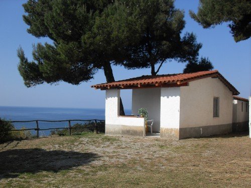 Casetta bianca con tetto rosso, immersa nel verde, vista mare e alberi circostanti.