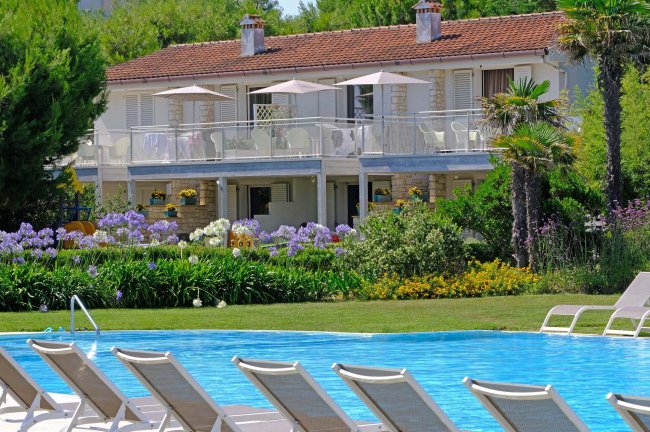 Villa con piscine, giardino fiorito, balconi e sdraio bianche in primo piano.