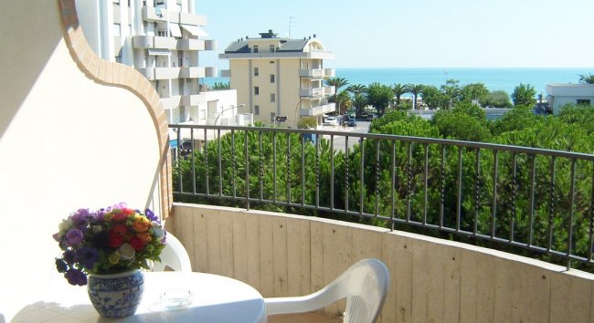 Un balcone con tavolo e fiori offre una vista sul mare e su edifici moderni.