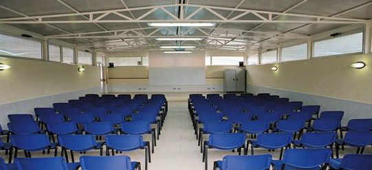 Aula spaziosa con sedie blu rivolte verso uno schermo bianco.