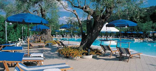 Area piscina con sdraio, ombrelloni blu e alberi intorno.