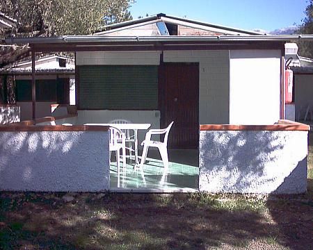 Bungalow con veranda e sedie bianche in plastica, circondato da un muretto in un giardino.