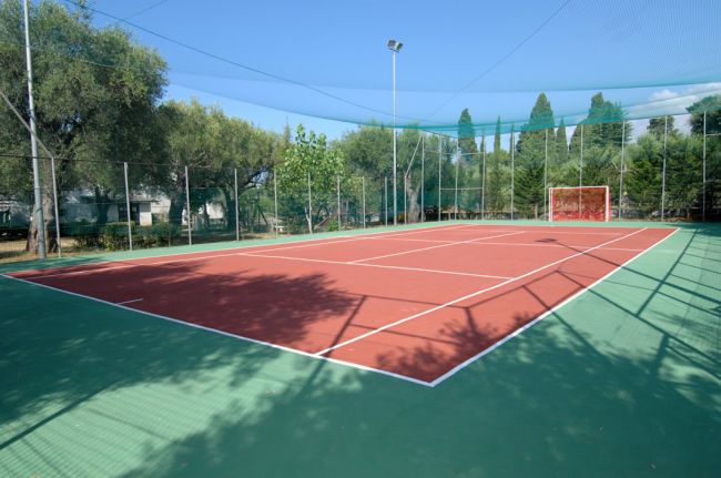 Campo sportivo all'aperto con superficie rossa, reti di protezione e alberi intorno.