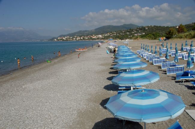 Una spiaggia con ombrelloni azzurri allineati e persone che nuotano nel mare.