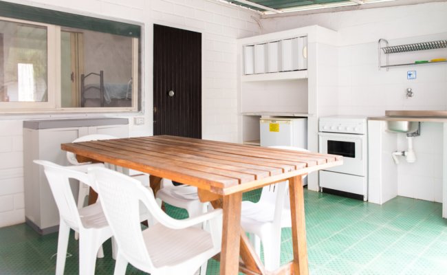 Cucina all'aperto con tavolo in legno, sedie bianche, frigorifero e lavello su pavimento verde.