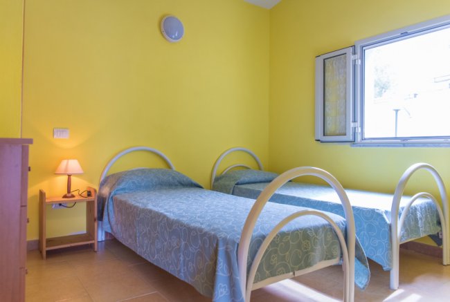 Due letti singoli con copriletti blu, parete gialla e finestra aperta.