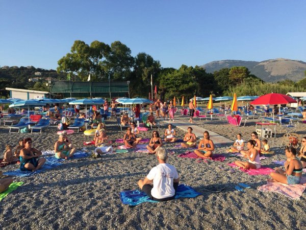Persone fanno yoga su una spiaggia, circondate da ombrelloni e alberi.