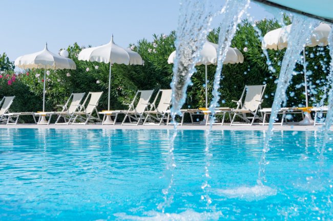 Dettaglio di getti d'acqua in una piscina, con lettini e ombrelloni bianchi sullo sfondo.