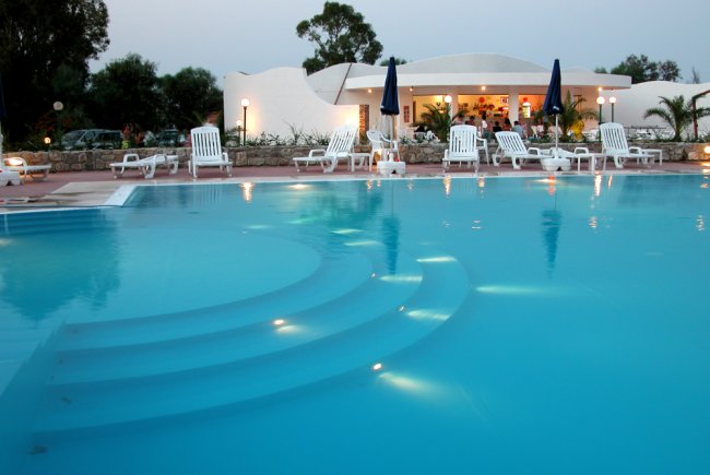 Piscina al tramonto con lettini bianchi e un bar sullo sfondo.