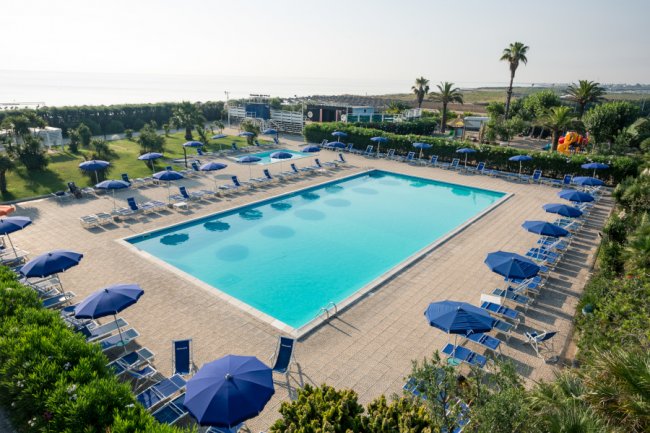 Piscina all'aperto con lettini e ombrelloni blu intorno, vista sul mare in lontananza.